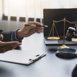 Two people in suits sit at a desk with a clipboard, golden scales of justice, and a judge's gavel, discussing how Business Formation & Tax Planning Lawyers set startups up for success in a well-lit office.