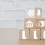 Four wooden blocks form a pyramid. Icons for a target, rocket, bank, and money highlight how business formation & tax planning lawyers set startups up for success. At the base, glowing "LLC" letters rest on a light wood surface with a white brick wall background.