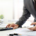 A person in a business suit uses a calculator and holds a smartphone while standing at a desk with documents, a laptop, and office supplies, preparing Year-End Tax Planning Tips for California Businesses.