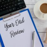 A clipboard with a paper titled "Year End Review," a pen, coffee, an hourglass, and part of a keyboard on a white wooden table—perfect for tackling U.S. Tax Compliance for Expats, Remote Workers & Travelers.