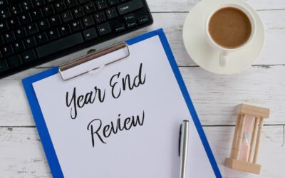 A clipboard with a paper titled "Year End Review," a pen, coffee, an hourglass, and part of a keyboard on a white wooden table—perfect for tackling U.S. Tax Compliance for Expats, Remote Workers & Travelers.
