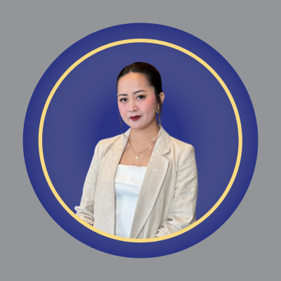 A woman with dark hair tied back wears a white top and light beige blazer. She stands against a blue background framed by a gold and blue circular border. A woman with dark hair tied back wears a white top and light beige blazer. She stands against a blue background framed by a gold and blue circular border.