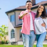 A smiling couple stands in front of their new house on a sunny day. The man holds keys toward the camera, celebrating homeownership and the benefits like the $40,000 SALT Deduction. Both are casually dressed and look happy and excited.