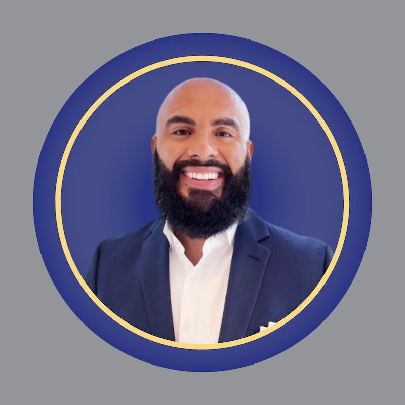 A smiling man with a bald head and full beard, wearing a dark suit jacket and white shirt, is centered in a circular frame with a blue background and yellow border. A smiling man with a bald head and full beard, wearing a dark suit jacket and white shirt, is centered in a circular frame with a blue background and yellow border.