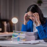 A woman in a blue blazer sits at a desk, looking stressed while holding her head. In front of her is a large stack of paperwork with colorful tabs, next to a laptop and calculator in an office setting.
