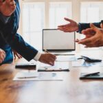 Four people in business attire discuss how to complete IRS Form 433-A around a wooden conference table, gesturing over charts, papers, a calculator, and a laptop. Only their hands and torsos are visible during the focused meeting.