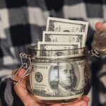 A person in a plaid shirt holds an open glass jar filled with several rolled and folded U.S. hundred-dollar bills, highlighting the potential benefits of No Tax on Tips in 2026.