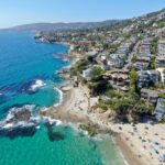 Aerial view of a coastal town with houses on hills overlooking a sandy beach and turquoise ocean, where people sunbathe and swim; perfect for a Tax Attorney in Irvine seeking relaxation by rocky outcrops under a clear blue sky.