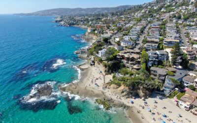 Aerial view of a coastal town with houses on hills overlooking a sandy beach and turquoise ocean, where people sunbathe and swim; perfect for a Tax Attorney in Irvine seeking relaxation by rocky outcrops under a clear blue sky.