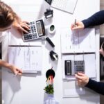 Two people sit across from each other at a desk, preparing for the 2026 Tax Season Kickoff with calculators, documents in binders, and pens. Office supplies, a plant, and a laptop are also spread across the white desk, viewed from above.