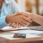 Two people shake hands across a desk with documents, a notebook, and a smartphone, suggesting a successful business agreement—perhaps after consulting an Offer in Compromise Guide. Both individuals wear long-sleeved shirts.