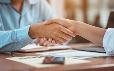 Two people shake hands across a desk with documents, a notebook, and a smartphone, suggesting a successful business agreement—perhaps after consulting an Offer in Compromise Guide. Both individuals wear long-sleeved shirts.