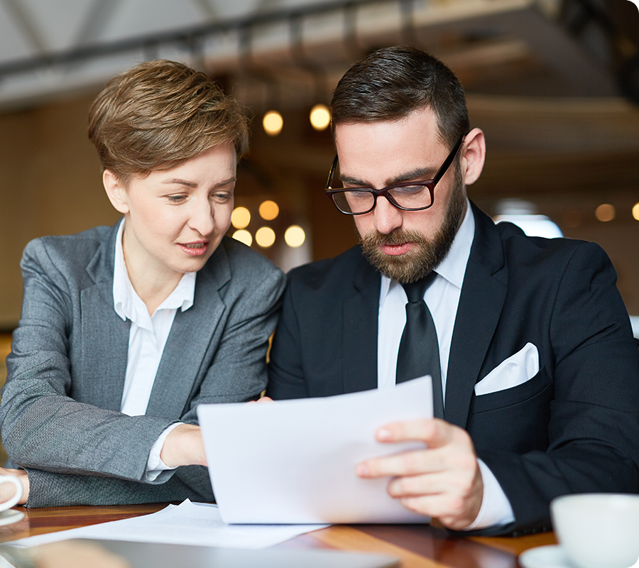 Two business professionals in formal attire sit together at a table, reviewing documents. The woman is leaning in and appears to be discussing the papers with the man, who is attentively reading them.