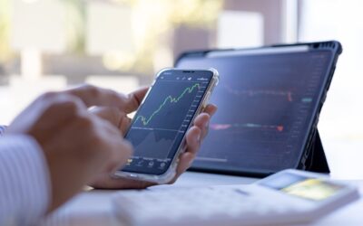 A person checks a smartphone displaying a rising stock chart; a tablet with financial graphs and a calculator are on the desk—highlighting the impact of Stock Option Taxes in California.