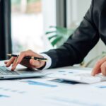 A person in a suit working at a desk, one hand on a laptop and the other pressing calculator buttons—focused on California FTB Audit Defense, with charts, graphs on paper, and a small potted plant in the background.