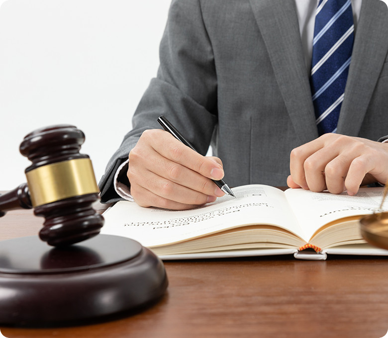 A person in a gray suit and blue striped tie is writing in a book at a desk, with a judge's gavel and scales of justice visible in the foreground.