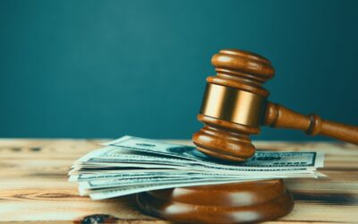 A judge’s gavel rests on a wooden block with a stack of US dollar bills underneath, symbolizing legal decisions and FBAR Penalties 2026, all set on a wooden table against a teal background.