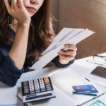 A woman sits at a desk holding envelopes, appearing concerned while on the phone. Surrounded by documents and charts, she seems to be researching how to stop an IRS bank levy in California as she manages her finances.