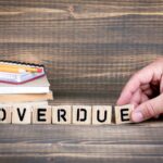 A hand arranges wooden blocks spelling "OVERDUE." In the background, there are stacked notebooks and pencils on a wooden surface, hinting at missed paperwork ahead of the Tax Extension Deadline 2026.
