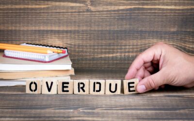 A hand arranges wooden blocks spelling "OVERDUE." In the background, there are stacked notebooks and pencils on a wooden surface, hinting at missed paperwork ahead of the Tax Extension Deadline 2026.