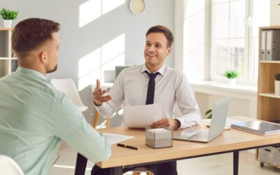 Two men sitting across from each other at a desk in a bright office. One man is smiling and holding papers, appearing to interview or have a discussion with the other man, who listens attentively. A laptop is on the desk.