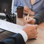 A person hands a clipboard with a document to another person, who is signing it. A laptop, pencil holder, and coffee cup are on the table, and two people sit across from the person with the clipboard.