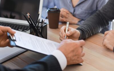 A person hands a clipboard with a document to another person, who is signing it. A laptop, pencil holder, and coffee cup are on the table, and two people sit across from the person with the clipboard.