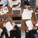 A group of professionals working together at a wooden table with laptops, tablets, papers, charts, notebooks, coffee cups, and pens, discussing data and collaborating on a project.