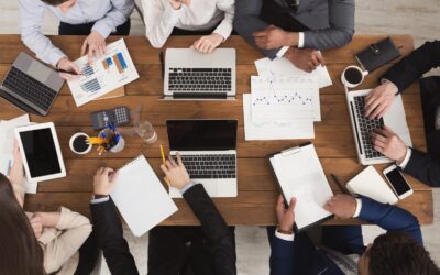 A group of professionals working together at a wooden table with laptops, tablets, papers, charts, notebooks, coffee cups, and pens, discussing data and collaborating on a project.
