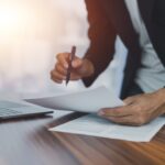 A person in a suit holding papers and a pen, leaning over a desk with a laptop, reviewing documents in a bright office setting.