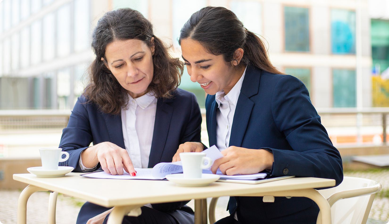 Two women in business suits sit at an outdoor table with coffee cups, smiling and looking at documents together, appearing to discuss work or study materials.
