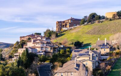 Houses with tan exteriors line a winding road on a green hillside under a blue sky, with larger buildings and a round structure visible at the top of the hill. Trees and shrubs surround the homes.