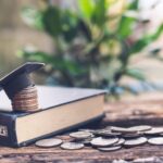 A graduation cap sits atop a stack of coins on a closed book, with more scattered coins on a wooden surface. A blurred green plant is in the background.