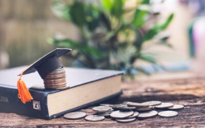 A graduation cap sits atop a stack of coins on a closed book, with more scattered coins on a wooden surface. A blurred green plant is in the background.