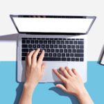 Top view of hands typing on a laptop keyboard, with a calculator on the left, an open notebook, and a pen on the right, all on a white and blue background.