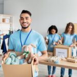 A smiling young man holds a box of donated items, including a stuffed bunny, while other volunteers sort clothes and supplies in the background at a donation center.