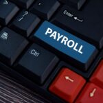 A close-up of a computer keyboard with a blue key labeled "PAYROLL" replacing the usual key, surrounded by standard black and red keys on a wooden surface.