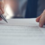 Close-up of a person’s hands holding a pen and signing a document on a desk, with text visible but out of focus.