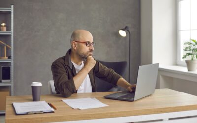 A man with glasses sits at a desk, looking thoughtfully at a laptop. Papers, a pen, and a coffee cup are on the desk. The room has a gray wall, a shelf, a lamp, and a window with natural light.