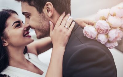 A happy couple embraces closely, smiling at each other. The woman’s hand, showing an engagement ring, rests on the man's neck. She holds a bouquet of pale pink roses behind his shoulder.