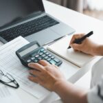 A person sits at a desk using a calculator, writing in a notebook. Nearby are financial documents, eyeglasses, and an open laptop. The scene suggests accounting, budgeting, or financial planning work.