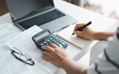 A person sits at a desk using a calculator, writing in a notebook. Nearby are financial documents, eyeglasses, and an open laptop. The scene suggests accounting, budgeting, or financial planning work.