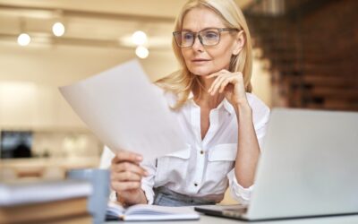 A woman wearing glasses and a white shirt sits at a desk with a laptop, thoughtfully looking at a document she is holding, with books and papers around her in a home office setting.
