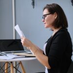 A woman wearing glasses sits at a desk with papers, a laptop, and stationery, reading a document. She appears focused and is dressed in business casual attire. The workspace has a modern, minimalist style.
