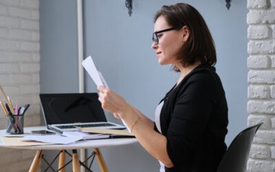 A woman wearing glasses sits at a desk with papers, a laptop, and stationery, reading a document. She appears focused and is dressed in business casual attire. The workspace has a modern, minimalist style.