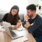 A woman and a man sit at a table with a laptop, papers, notebook, and calculator, looking at documents together as they appear to discuss finances or budgeting in a home setting.