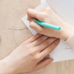 A person writes on lined paper with a turquoise pen at a wooden desk, beside an open notebook, colorful folders, and a smartphone.