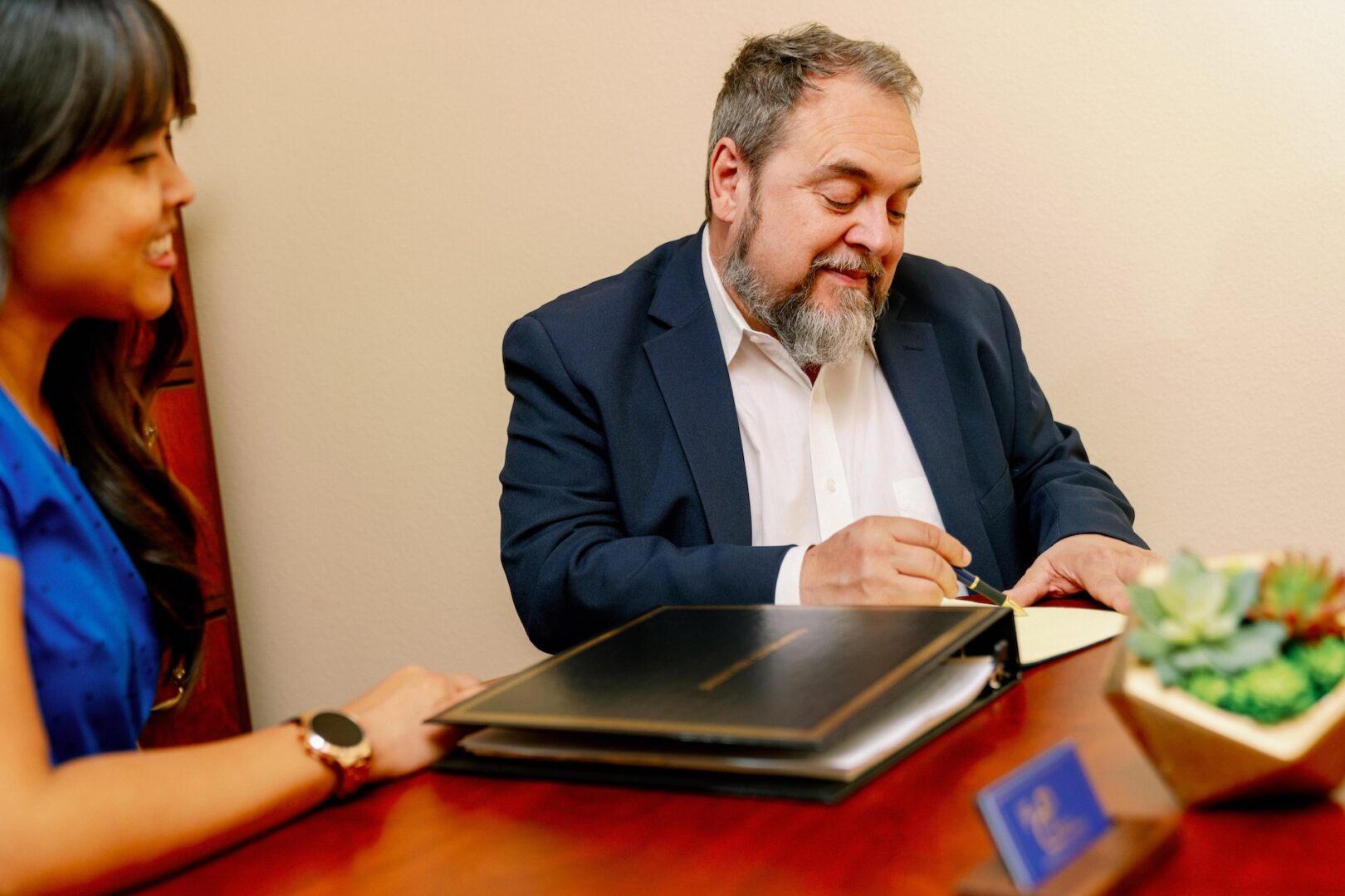 A man in a suit signs a document at a wooden desk while a woman in a blue dress sits beside him, smiling. A folder, a small plant, and a nameplate are on the desk. A man in a suit signs a document at a wooden desk while a woman in a blue dress sits beside him, smiling. A folder, a small plant, and a nameplate are on the desk.