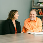 Two people sit at a desk, smiling and talking. The woman wears a black blazer, and the man wears an orange shirt. A notepad, plant, and bookshelf with red books and framed photos are visible in the background.