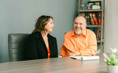 Two people sit at a desk, smiling and talking. The woman wears a black blazer, and the man wears an orange shirt. A notepad, plant, and bookshelf with red books and framed photos are visible in the background.